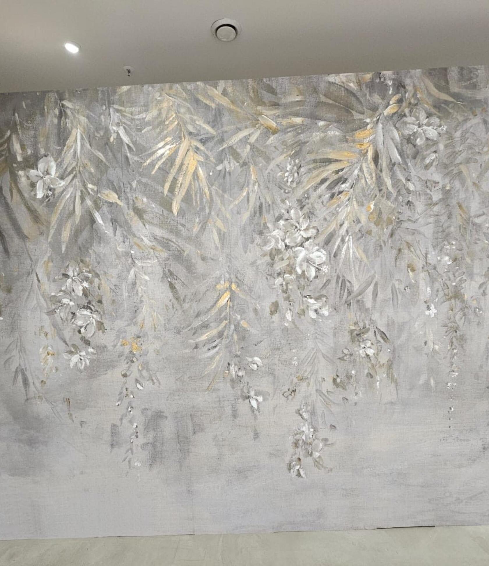 Großflächiges Wandbild mit zart gemalten hängenden Blättern und Blüten in Grautönen mit goldenen Akzenten, auf einer Innenwand unter Deckenleuchte, mit sichtbarem Bodenabschluss.