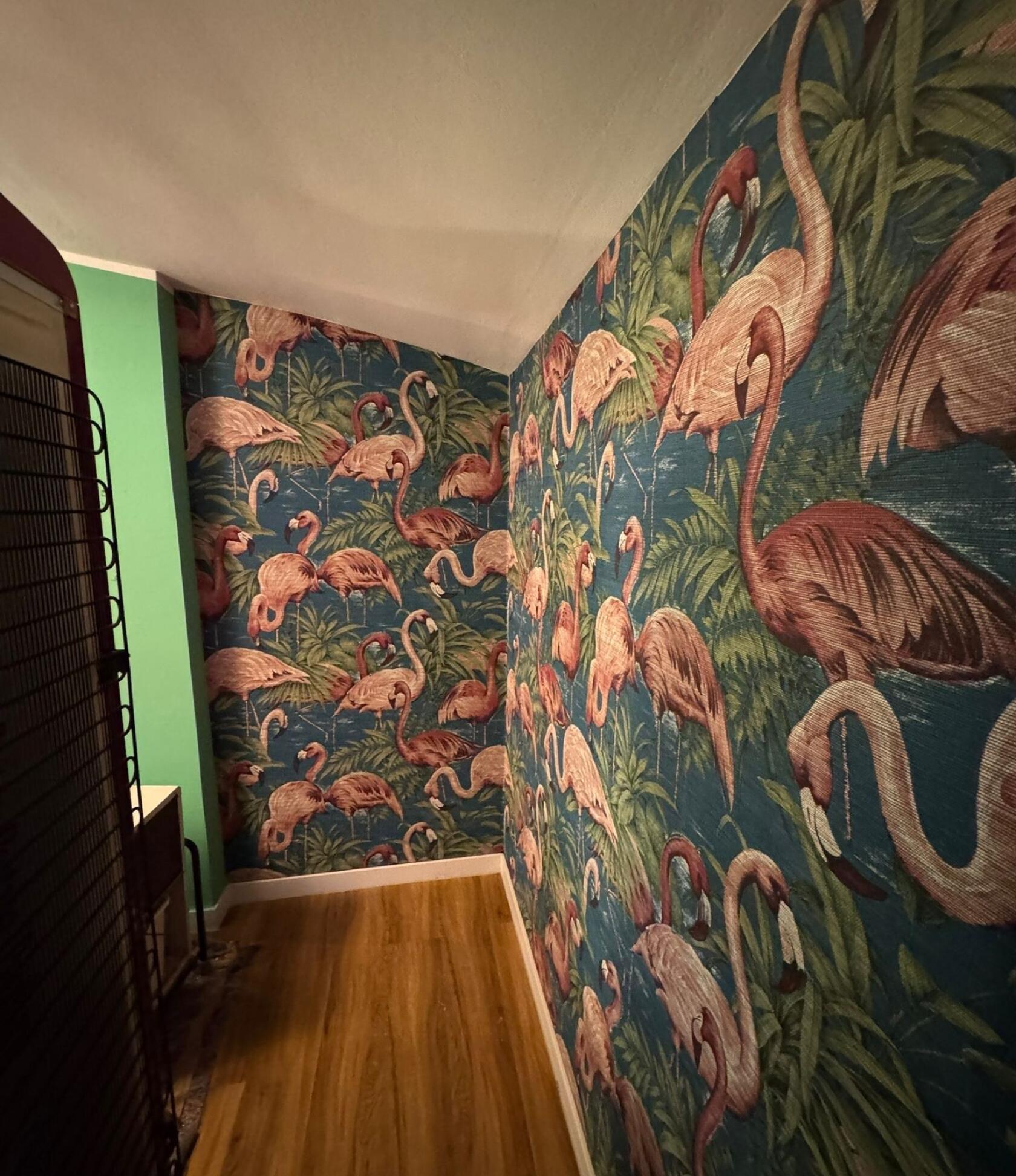 Tapete mit großem rosa Flamingo-Motiv, mehrere Flamingos stehen und biegen die Hälse, bedeckt zwei angrenzende Wände einer schmalen Ecke mit Holzboden und schräger Decke.
