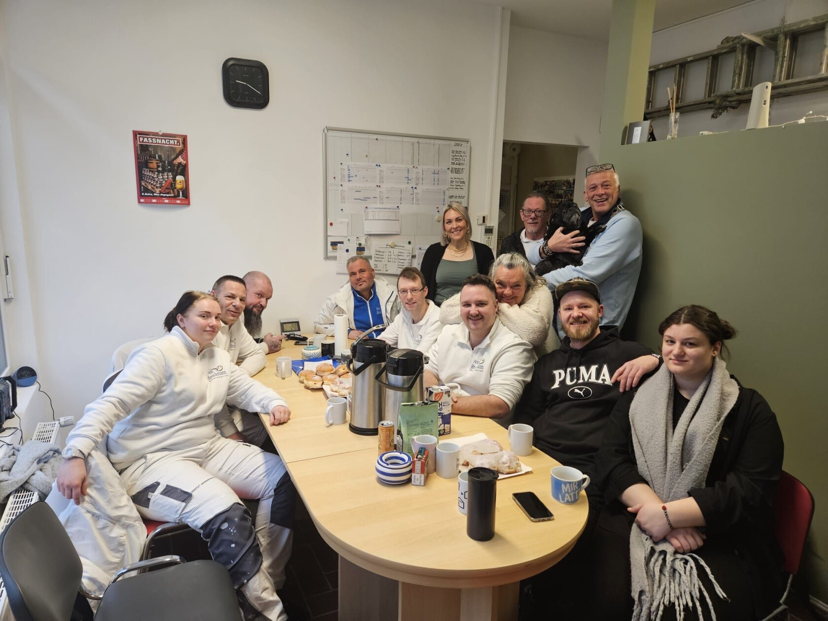 Mehrere Personen sitzen um einen Tisch mit Thermoskannen, Tassen und Gebäck; eine Person hält einen kleinen schwarzen Hund; im Hintergrund Whiteboard und Wandposter mit dem Text 