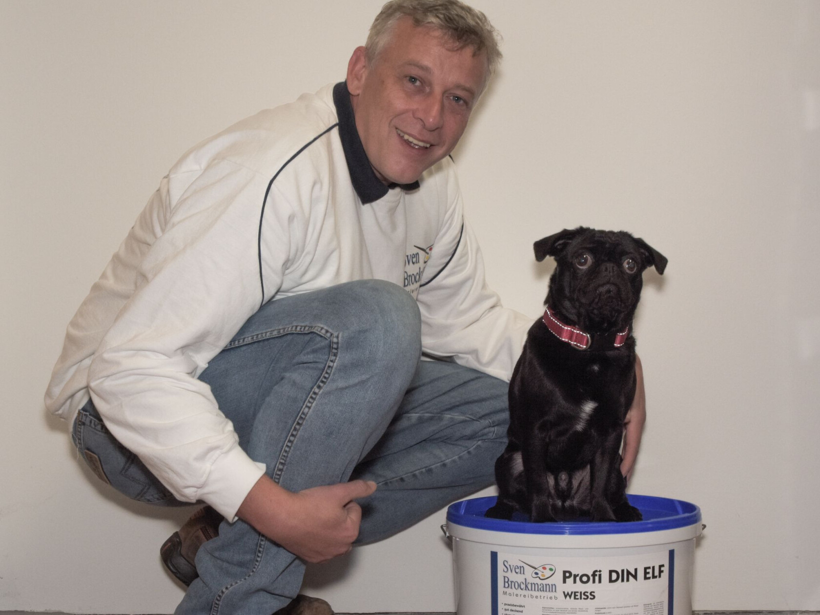 Schwarzer Mops sitzt auf einem weißen Farbeimer, ein hockender Mann in Jeans und weißem Pullover hält ihn neben einer weißen Wand. Text auf Eimer: 