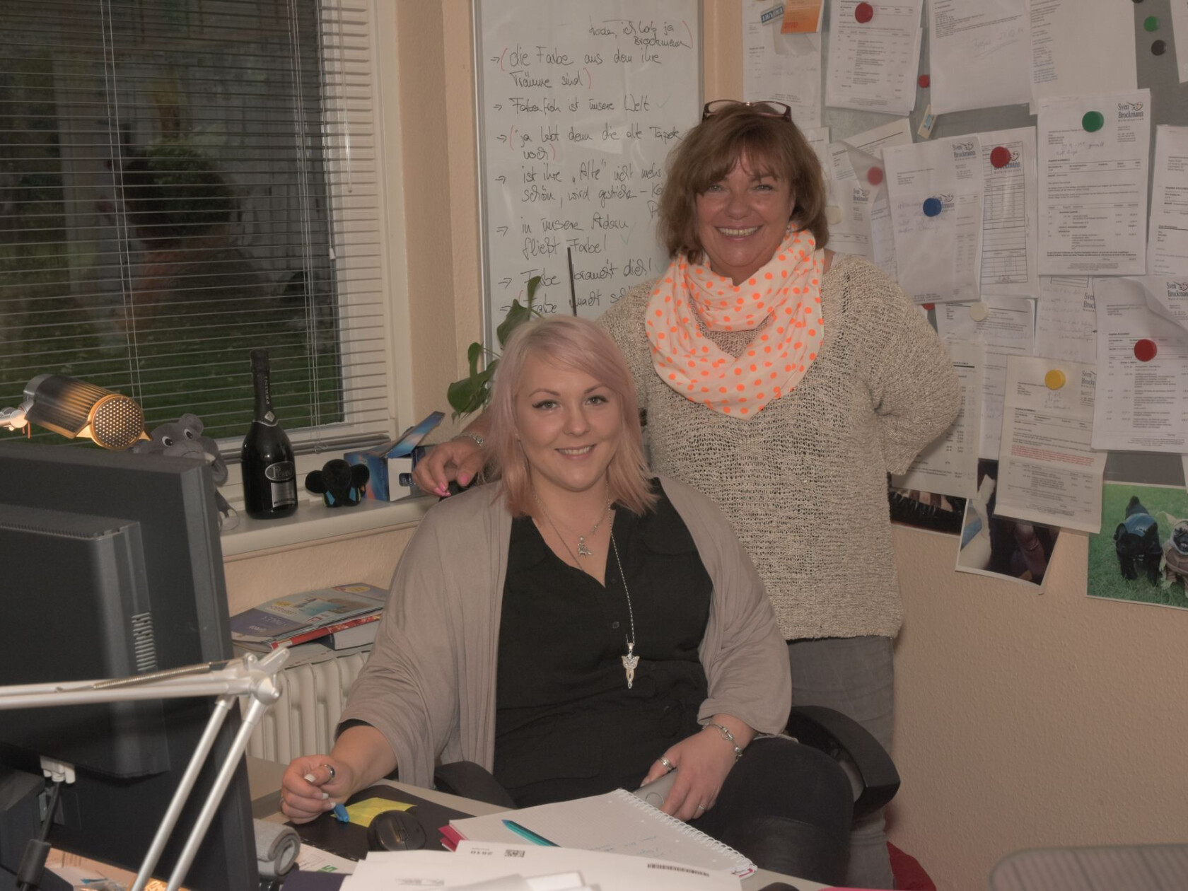 Zwei Frauen, eine sitzt am Schreibtisch mit Computer, die andere steht hinter ihr und legt Hand auf Stuhllehne; Büro mit Whiteboard, Pinnwand, Fenster und Büromaterial.