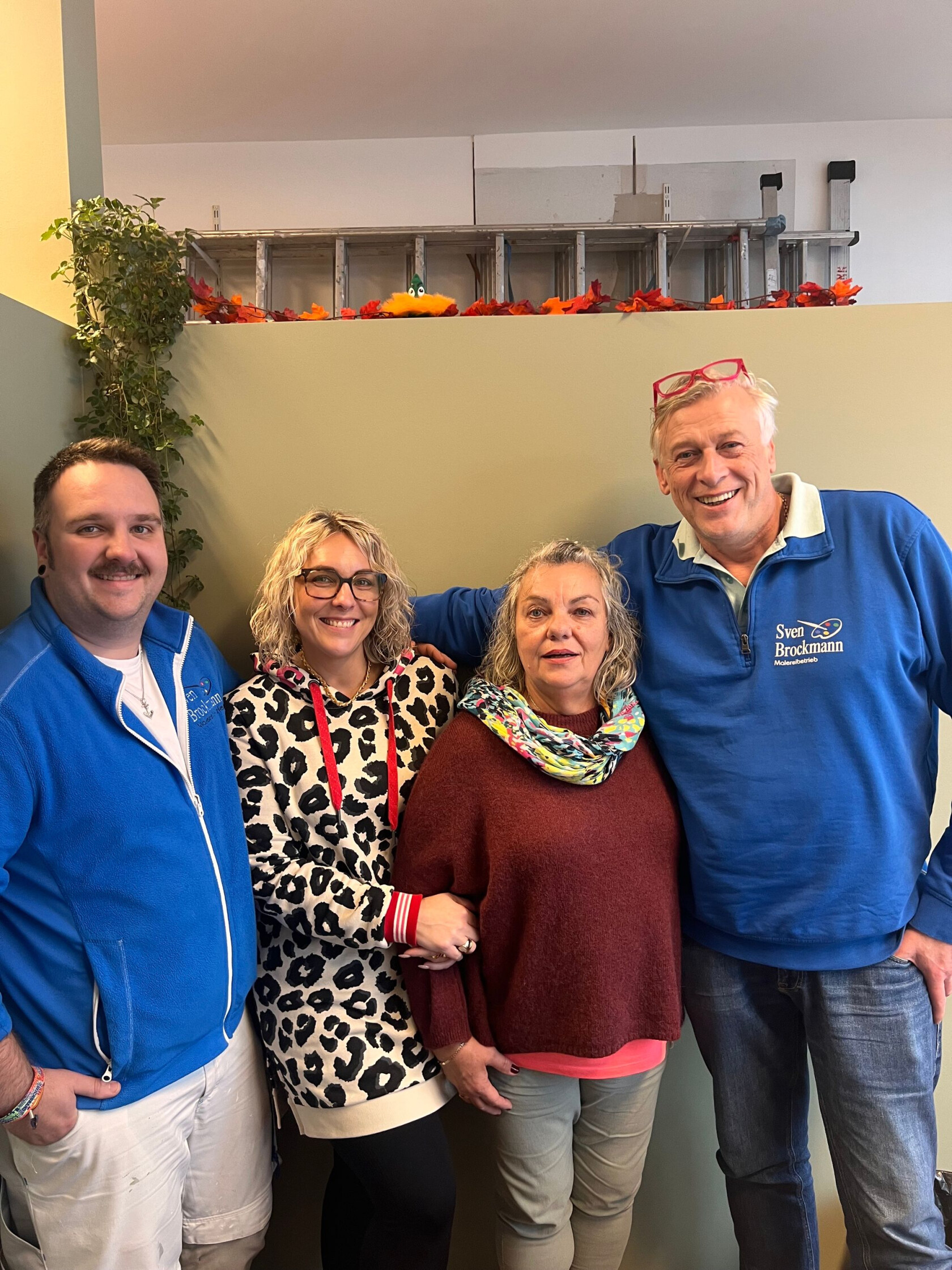 Vier Erwachsene stehen dicht nebeneinander und lächeln; zwei Männer und zwei Frauen, einer trägt ein blaues Shirt mit Text 