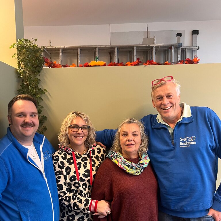 Vier Erwachsene stehen dicht nebeneinander und lächeln; zwei Männer und zwei Frauen, einer trägt ein blaues Shirt mit Text 