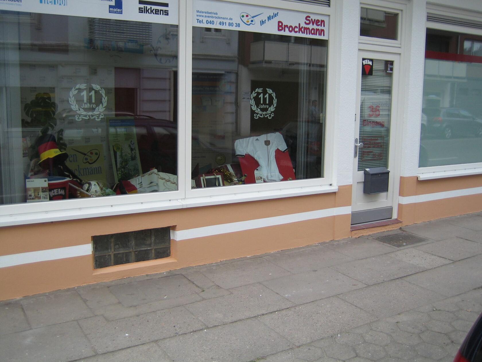Schaufenster eines Malerbetriebs, zeigt hinter Glas ein weißes Trikot, Trophäen, Fanartikel und Ausstellungsstücke; rechter Eingang mit Briefkasten und Gehweg davor. Sichtbare Schrift: 