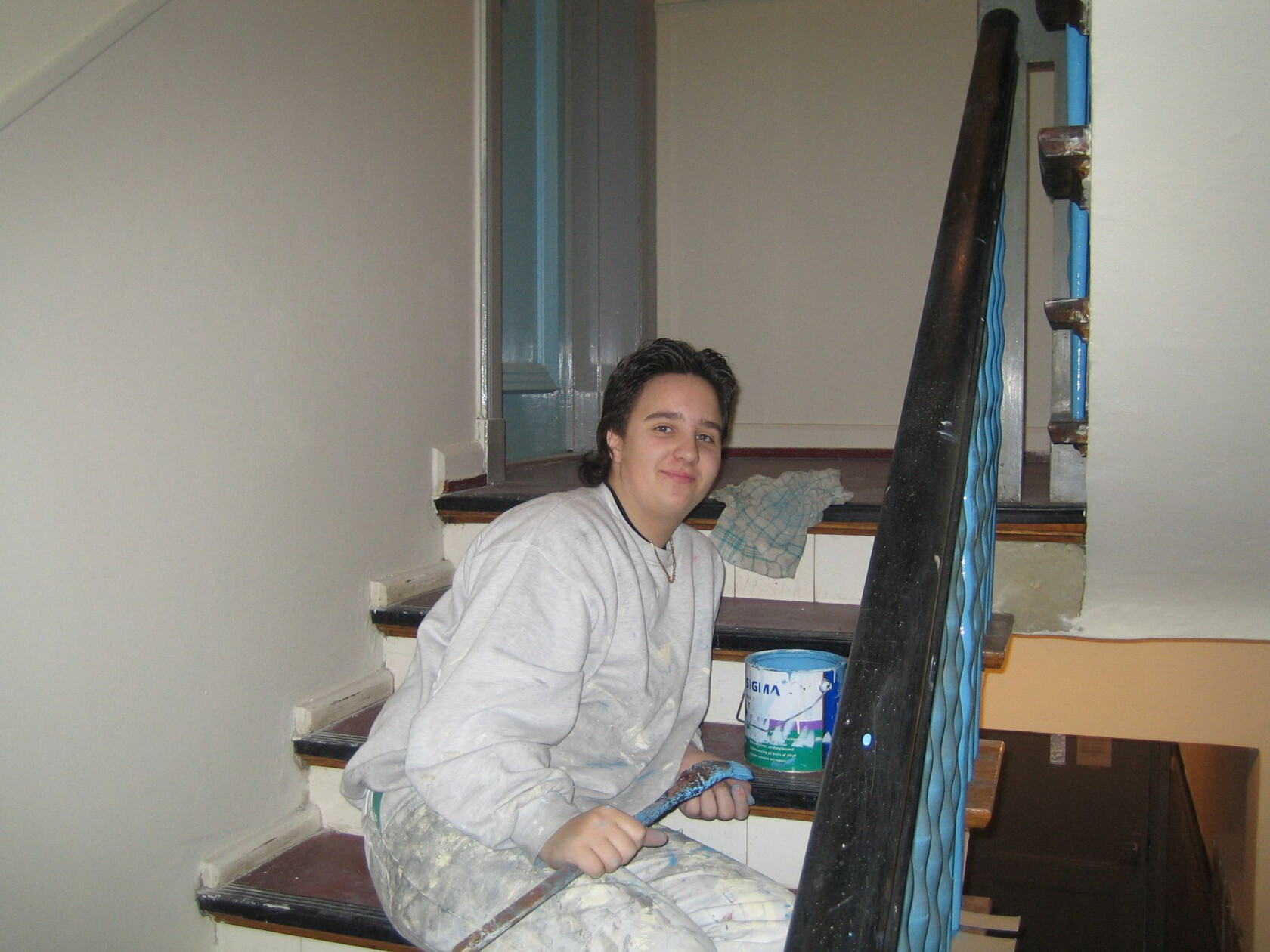 Person sitzt auf Treppenstufe und hält eine Farbrolle mit blauer Farbe; Innen-Treppenhaus mit schwarzem Handlauf, gedrehten blauen Geländerstäben, Farbeimer mit Aufschrift SIGMA und Lappen.