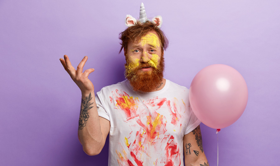 Image of puzzled displeased indignant man with ginger hair and beard, raises hand hesitantly, wears white t shirt dirty with paints, carries balloon, stands over purple background. Festive day